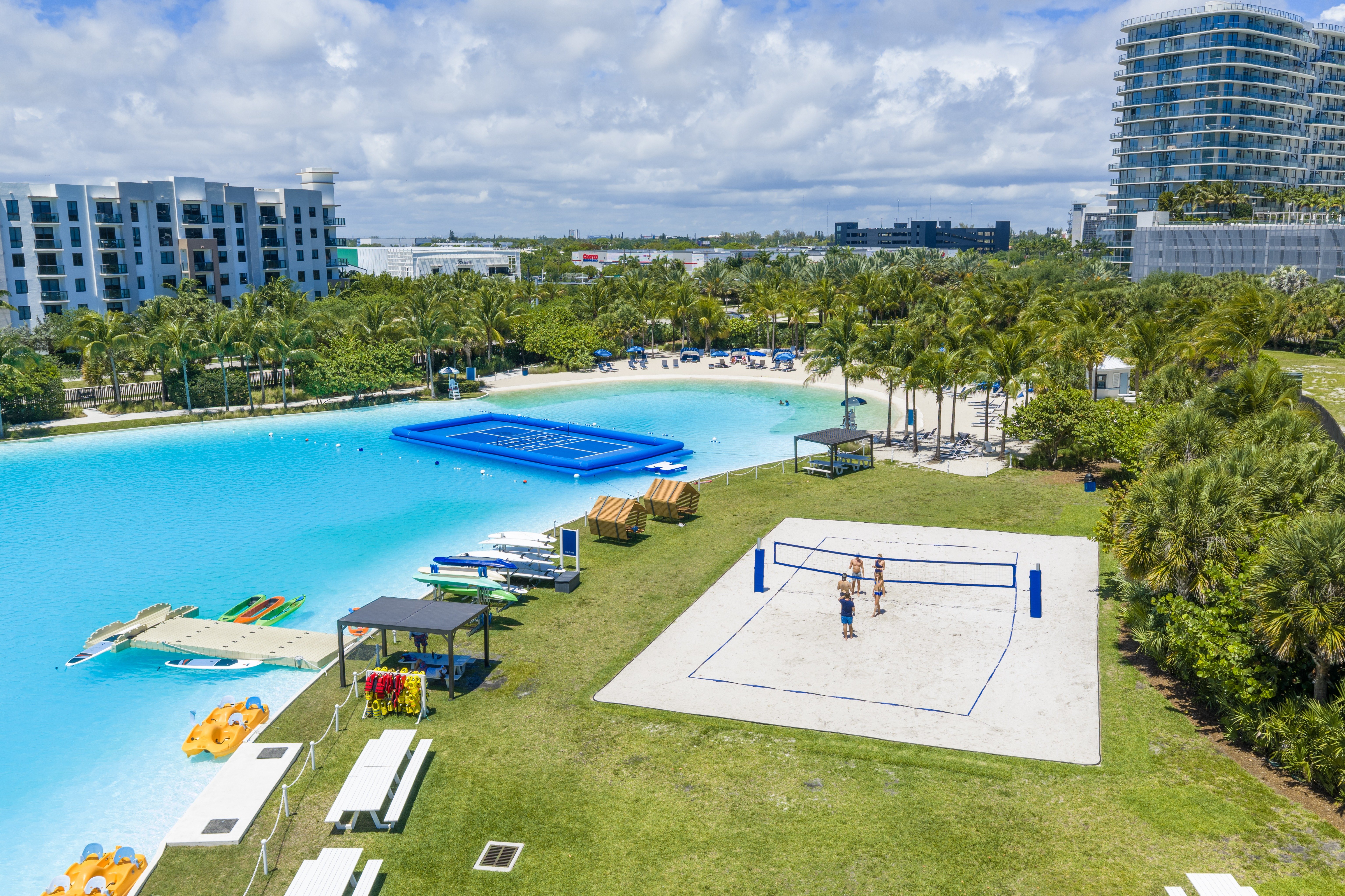 A beach volleyball court is surrounded by a pool and palm trees.